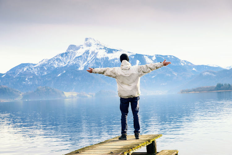 Man with arms outstretched standing on pier over lake against mountains