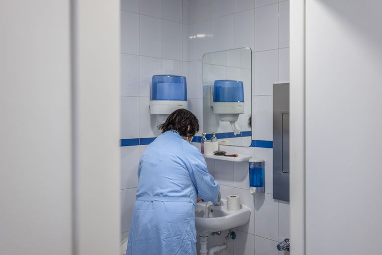Back view of anonymous elderly female in bathrobe washing hands in basin in restroom of hospital ward