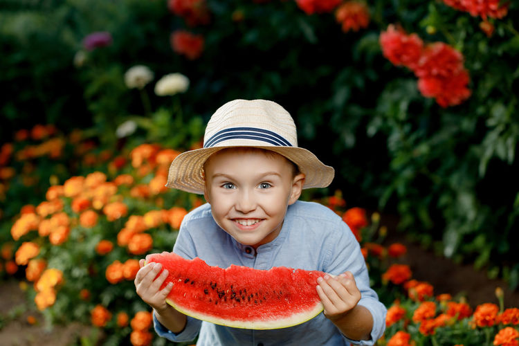 Shy little boy in a he holds a large slice of watermelon and hides his face outdoors. a cute boy in