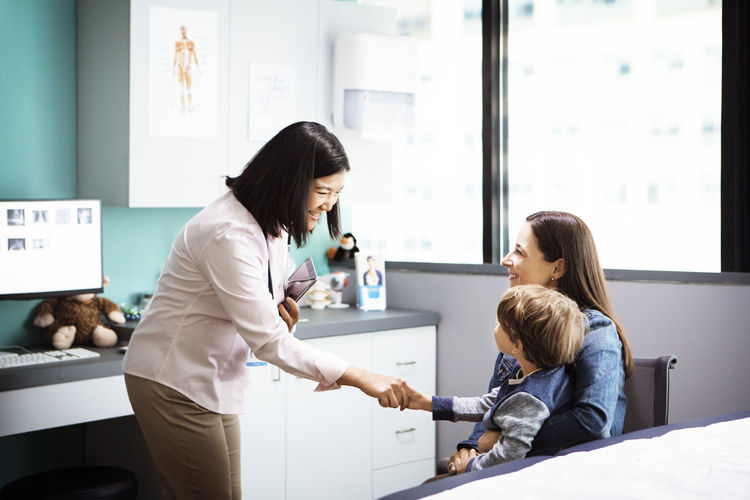 Happy female doctor greeting boy sitting with mother in clinic