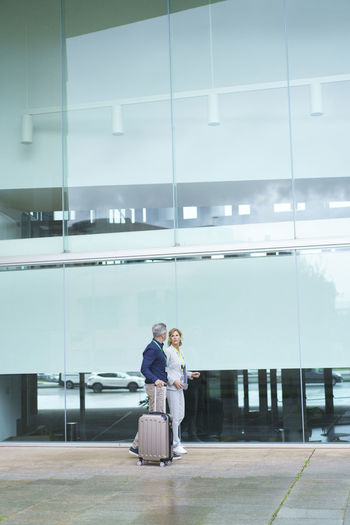 Businessman with suitcase walking by businesswoman in front of glass wall