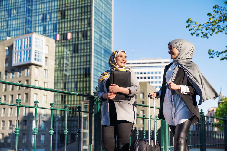Smiling muslim female entrepreneurs with suitcase walking along street in city and looking at each other