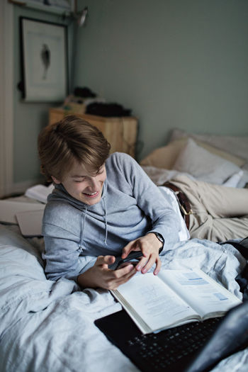Smiling teenage boy using social media on mobile phone while doing homework in bedroom