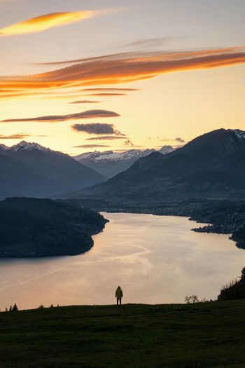 Rear view of person standing by lake against mountain range during sunset