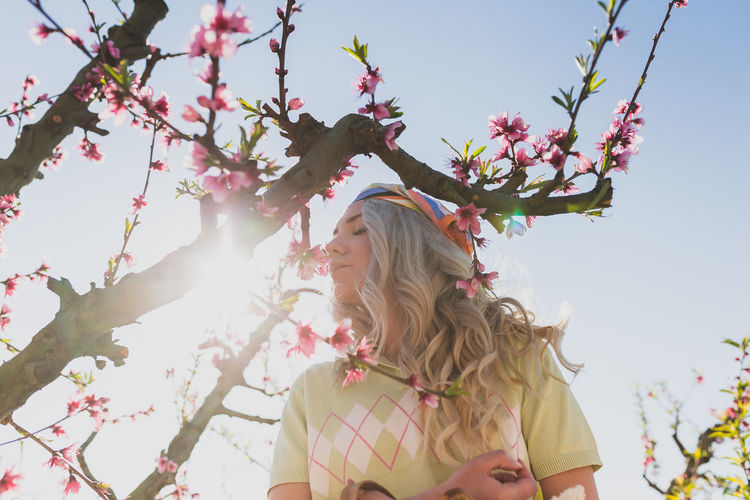 Low angle of female with closed eyes enjoying fragrance of fresh flowers growing on tree branches in garden