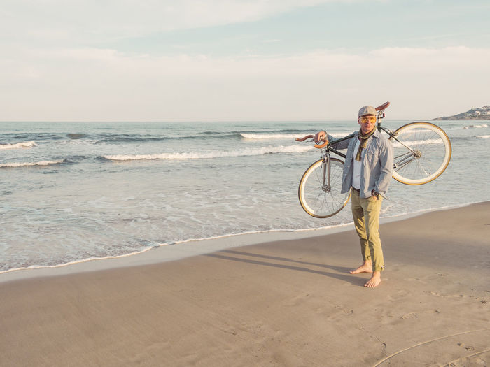 Barefoot man standing with fixie bike on his shoulder on the beach