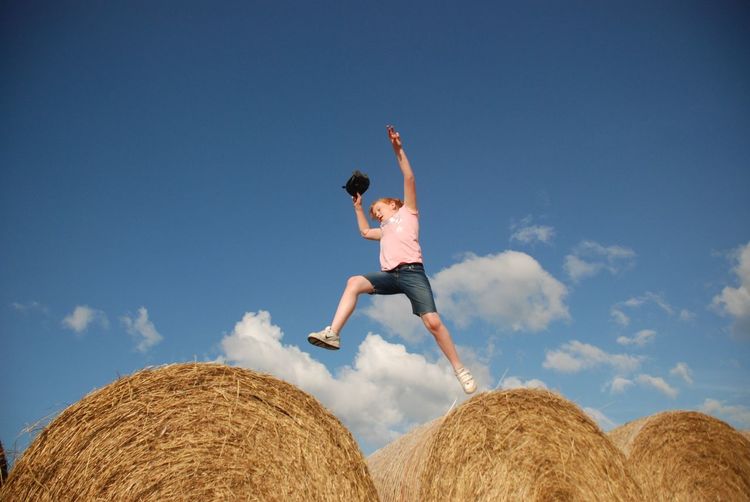 Low angle view of young woman jumping on hay bales against sky