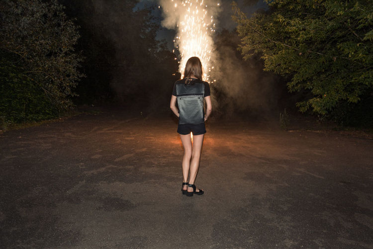 Rear view of young woman standing by illuminated firework on road at night