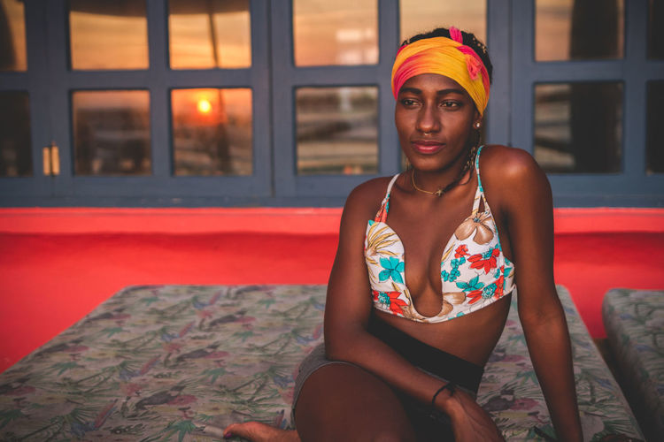 Pensive calm african american female wearing colorful bikini top and headband sitting on lounge in room with red floor and red sunset through window in background