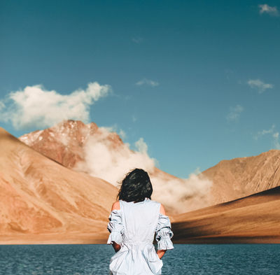 Rear view of woman standing by lake against mountain and sky