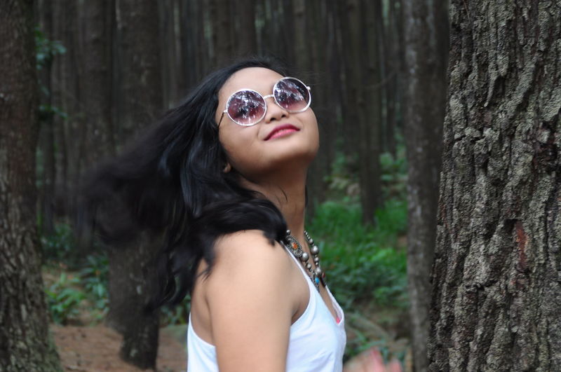 Smiling young woman wearing sunglasses while tossing hair at forest