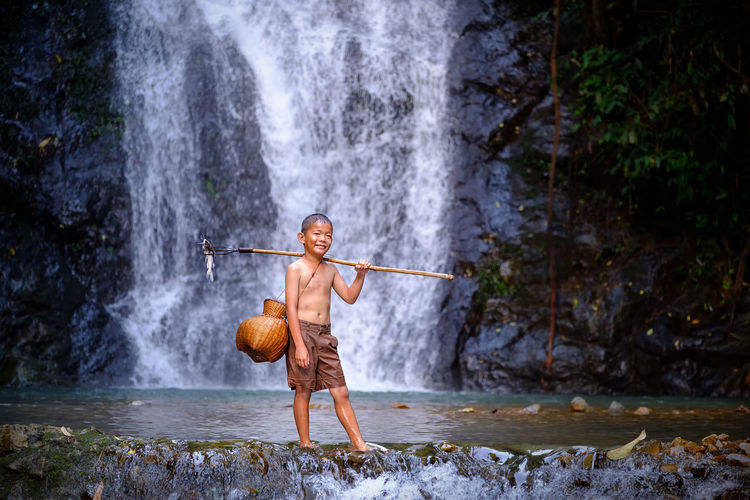 Full length of shirtless boy standing in river against waterfall