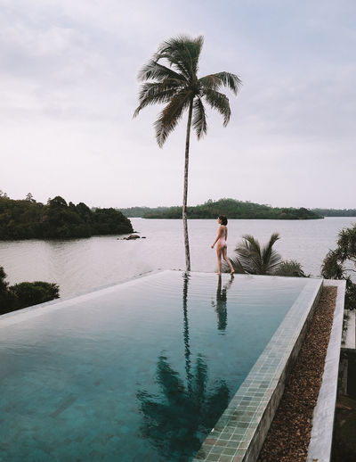 Side view of woman standing at edge of infinity pool against sky