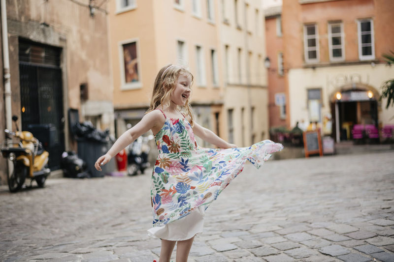 Happy cute girl wearing dress spinning on street against buildings in city