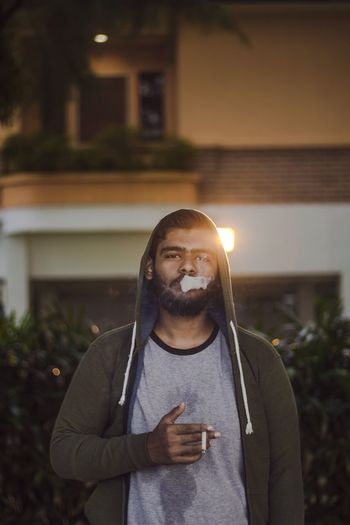 Portrait of young man smoking cigarette against building at night