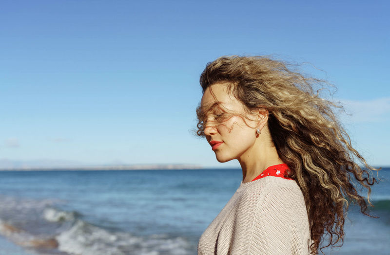 Side view of young woman with curly hair standing at beach against clear blue sky
