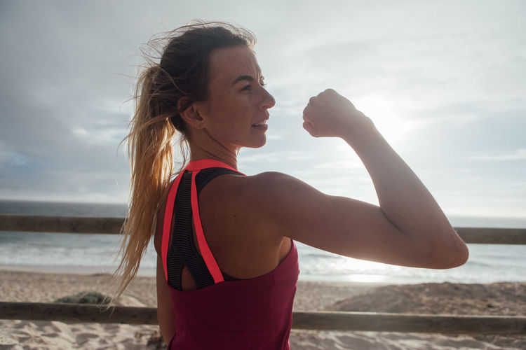 Woman flexing muscle at beach against sky