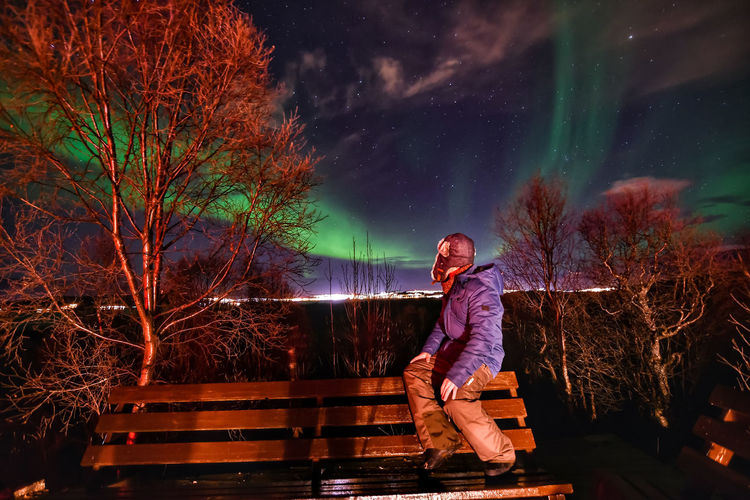 Side view of person sitting on bench against northern lights at night