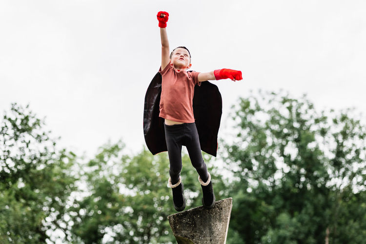 Boy in red superhero cape and hedgehog mask looking away while standing with arms raised with clenched fist pretending to fly on stone block on blurred background of park trees
