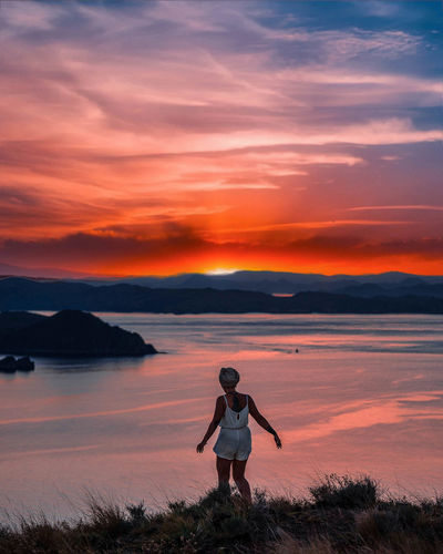 Rear view of woman walking on grass by sea against sky during sunset