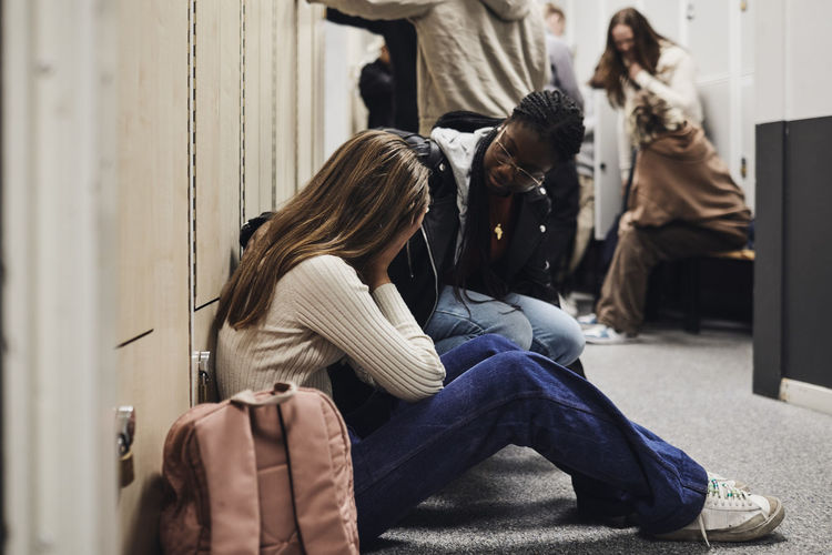 Teenage girl talking with depressed female friend sitting by backpack in school corridor