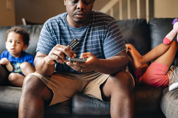 Father at home sitting on couch with kids in the background while holding tv remote and cellphone