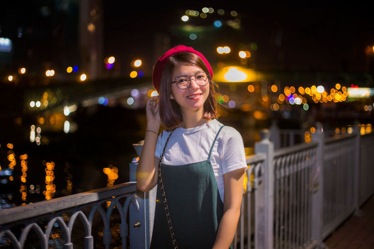 Portrait of young woman smiling while standing against canal in illuminated city at night