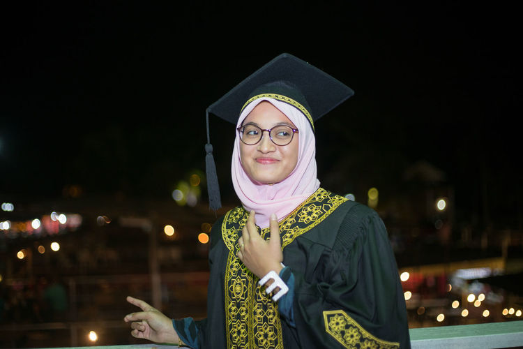 Smiling young woman wearing mortar board while standing against sky at night