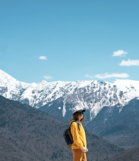 Young blond woman in yellow hoodie, cap, on background of snow capped caucasus mountains. hiking