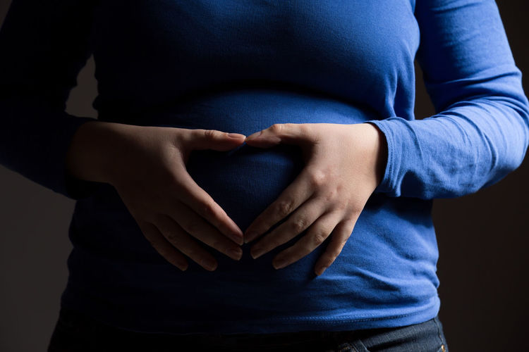 Midsection of pregnant woman making heart shape with hands against gray background