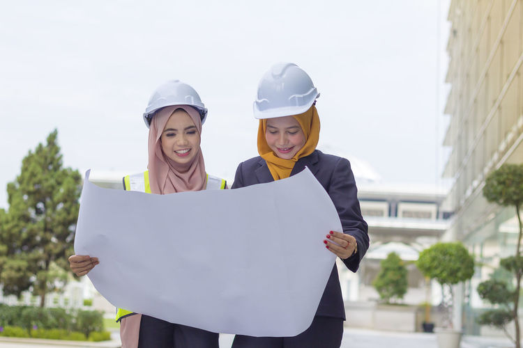 Happy female architects examining blueprint while standing against sky