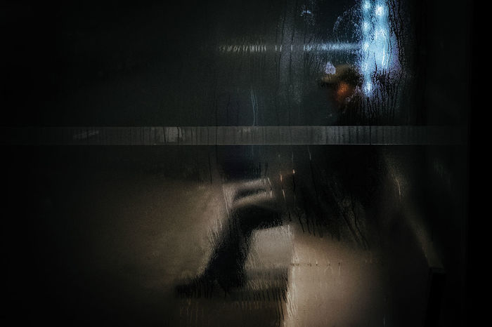 Man sitting on bench at night seen through wet glass window