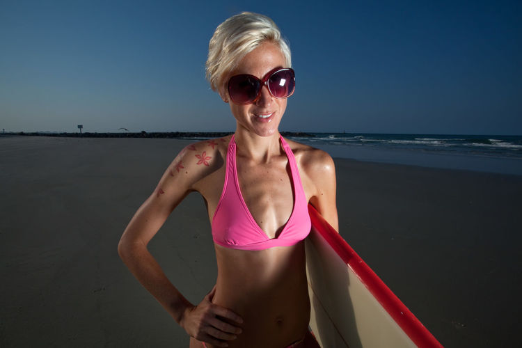 Portrait of smiling mid adult woman with surfboard standing at beach against sky