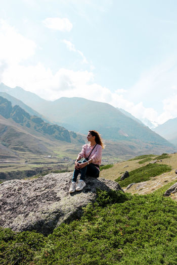 Full length of woman looking at view while sitting on rock against mountains