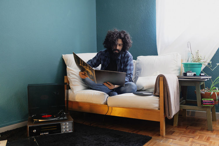 Bearded man with long hair reading record cover while sitting on sofa at home