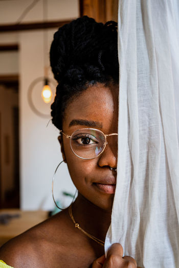 Pensive young african american woman in trendy eyeglasses covering half of face with curtain and looking at camera at home