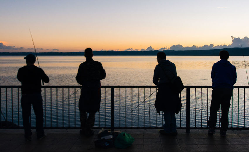 Rear view of friends fishing on pier over lake bracciano against sky during sunset