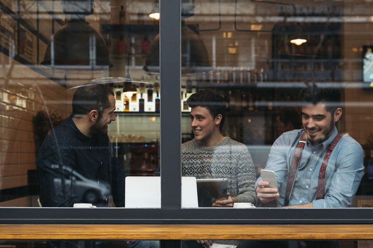 Friends working on laptop while sitting in cafe seen through window