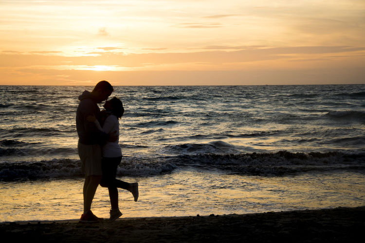Silhouette romantic couple standing on shore at beach against sky during sunset