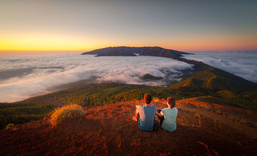 Back view of tender traveling couple sitting on hill while admiring spectacular scenery on clouds over mountainous terrain