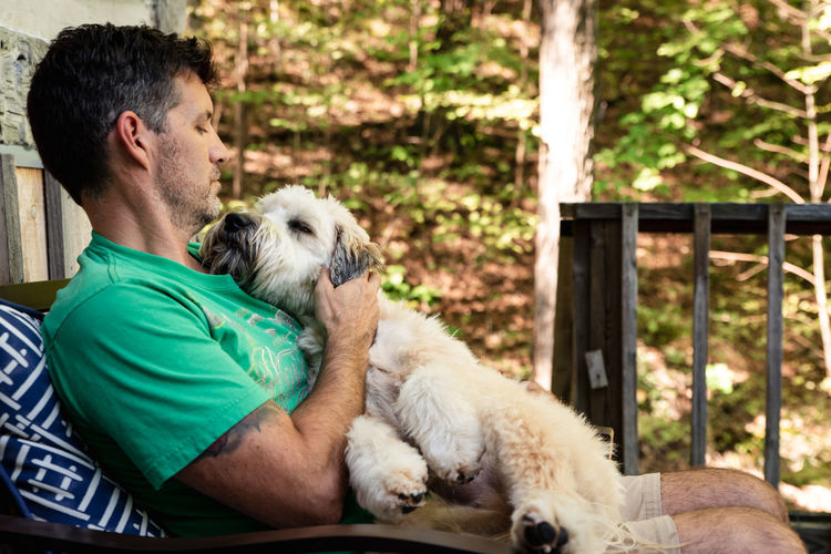 Man holding a fluffy dog on his lap outside on a deck in the woods.