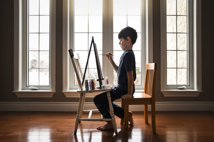 Young boy painting on a canvas on an easel in front of windows.