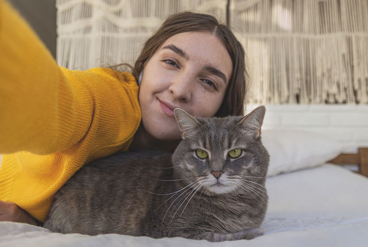 Happy woman in yellow sweater hugs her gray cat on the bed in a light interior