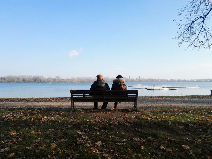 Romantica rear view of senior couple sitting on bench by lake against sky