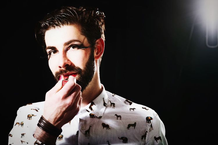 Portrait of man applying lipstick standing against black background