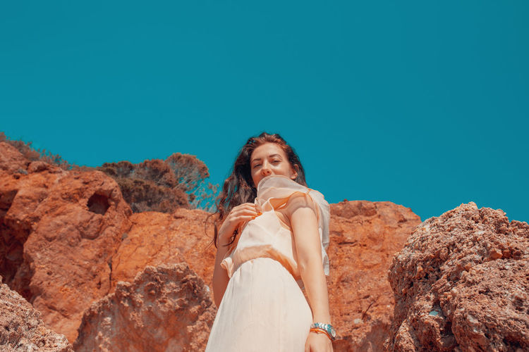 Low angle portrait of woman standing on rock against clear blue sky