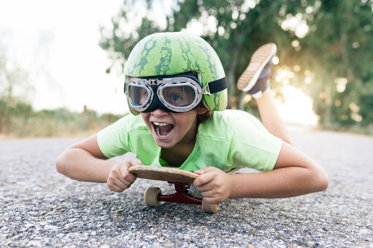 Ground level of happy kid in protective eyewear and ornamental watermelon helmet lying on skateboard on roadway and looking away