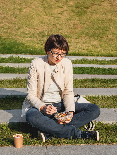 Woman sits on park bench with take away lunch box, coffee.  bowl with vegetables. healthy nutrition.