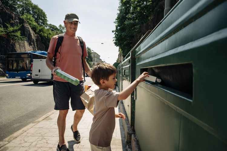 Boy trashing garbage in bin with grandfather at sidewalk