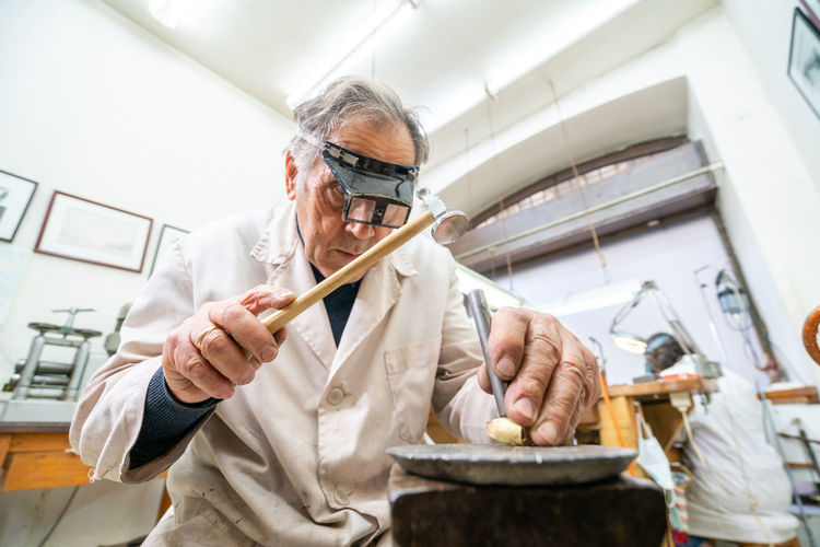 Low angle of focused skilled mature male master in glasses using hammer and chisel while working with gemstone in jewelry workshop